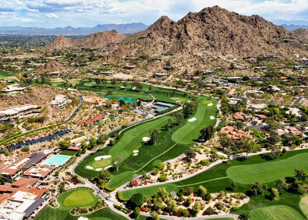 An aerial view of a desert landscape with a rugged mountain range. Surrounding the mountains are sprawling golf courses.