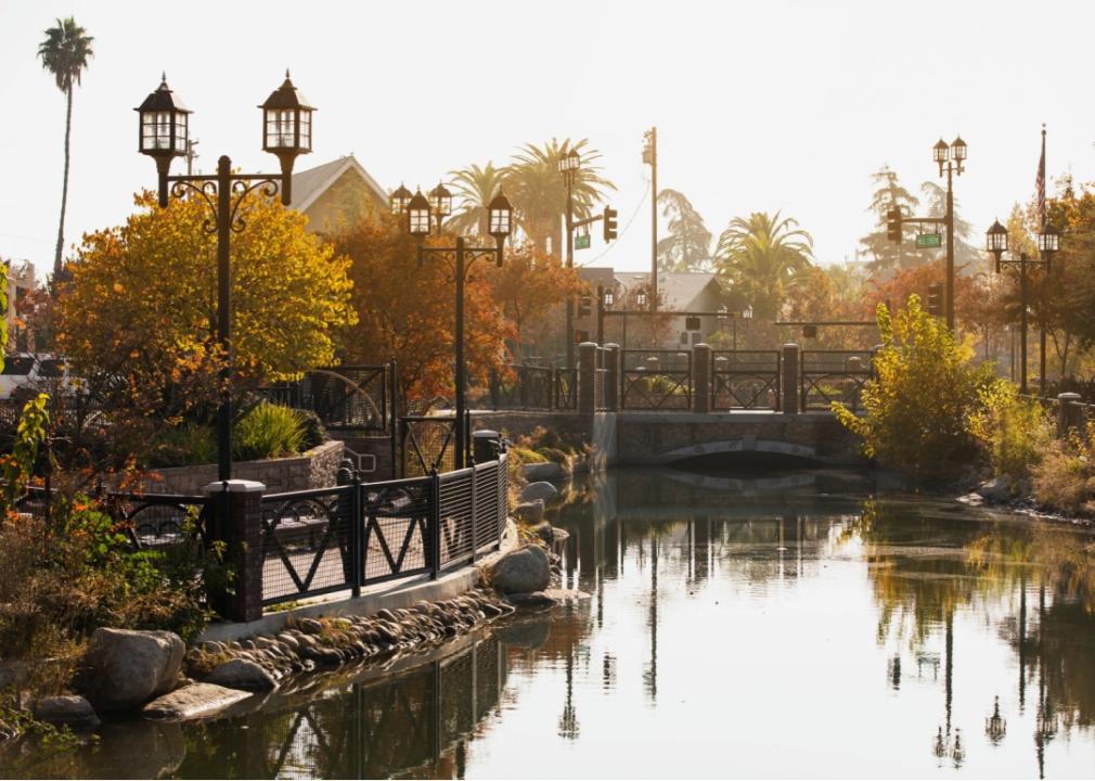 A park with a calm river flowing through it. A pedestrian bridge crosses the water, and the banks are lined with trees showing autumn foliage and decorative lampposts.