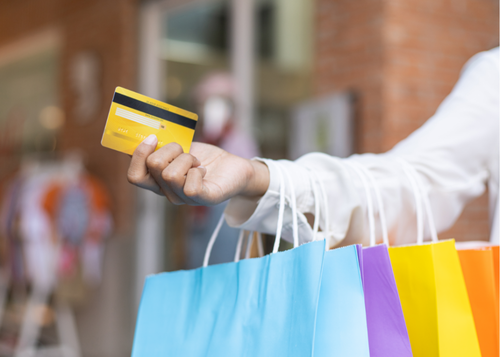 A woman's hand holding out a credit card while colorful shopping bags hang from her arm.