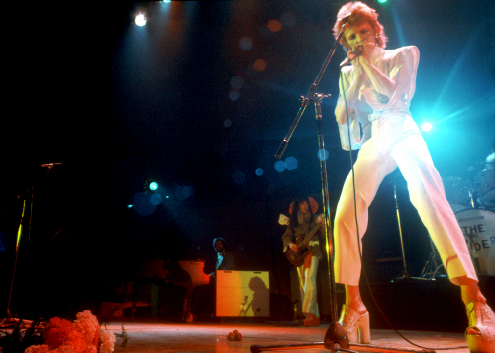 Bowie in a tight white suit with platform heels performs on stage. 