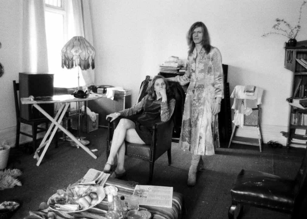 Angela Barnett sits in an arm chair with David Bowie in a dress standing beside her. 