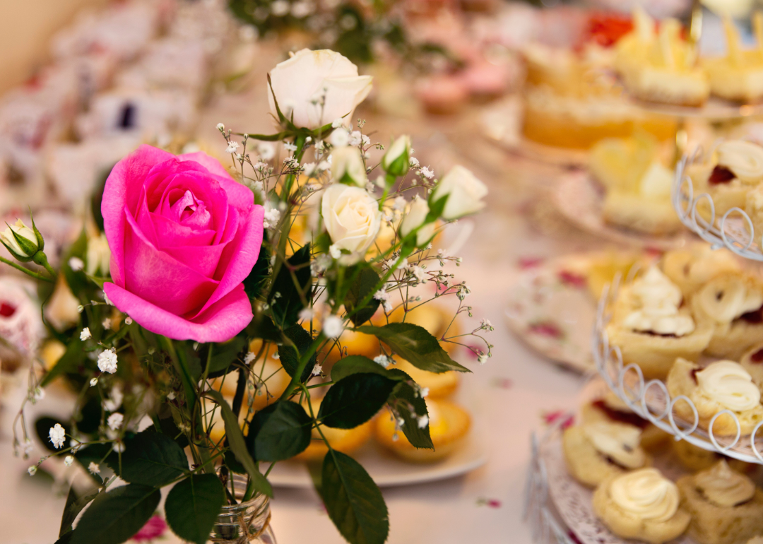 A single pink rose in front of a table spread with teacakes and pastries.