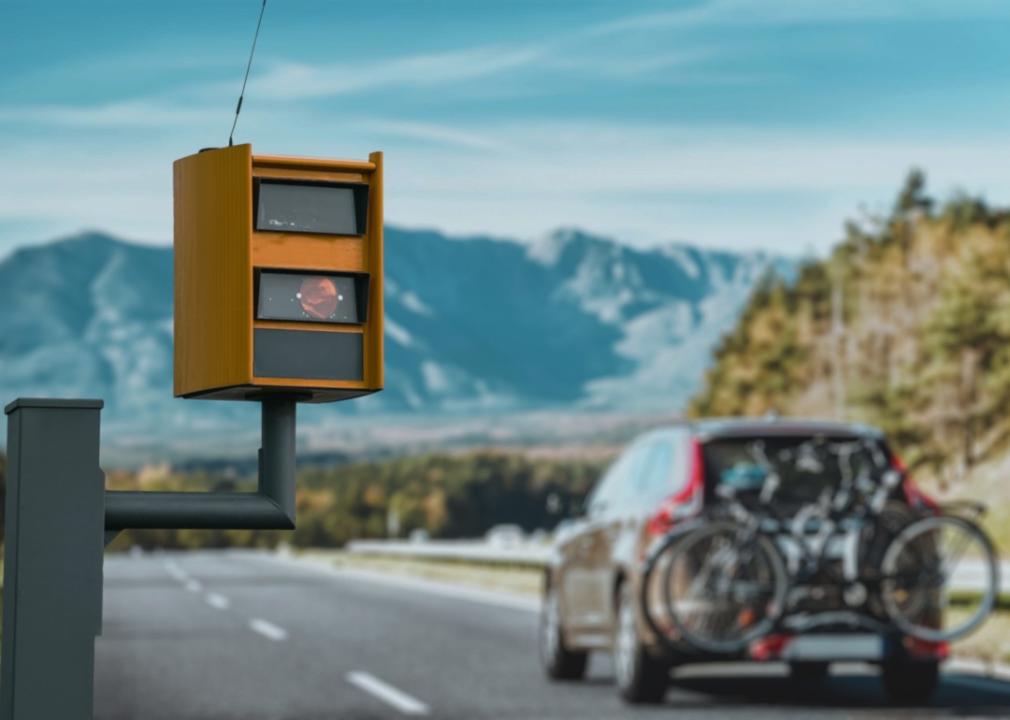 A yellow traffic speed camera prominently in the foreground, mounted on a pole along a highway. In the background, a car with bicycles mounted on its rear.