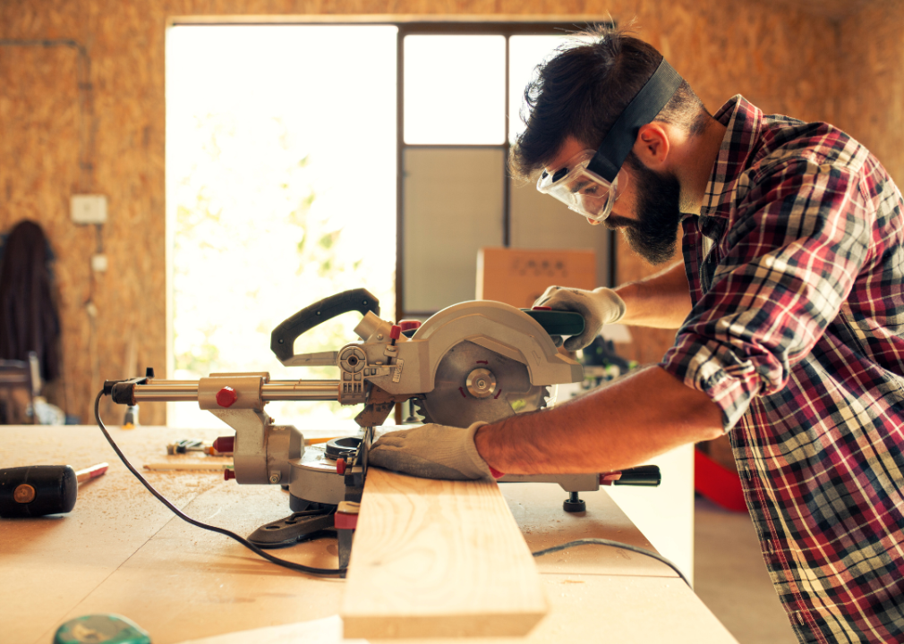 A man wearing eye protection mask holds a table saw in one hand and a long piece of wood in the other.