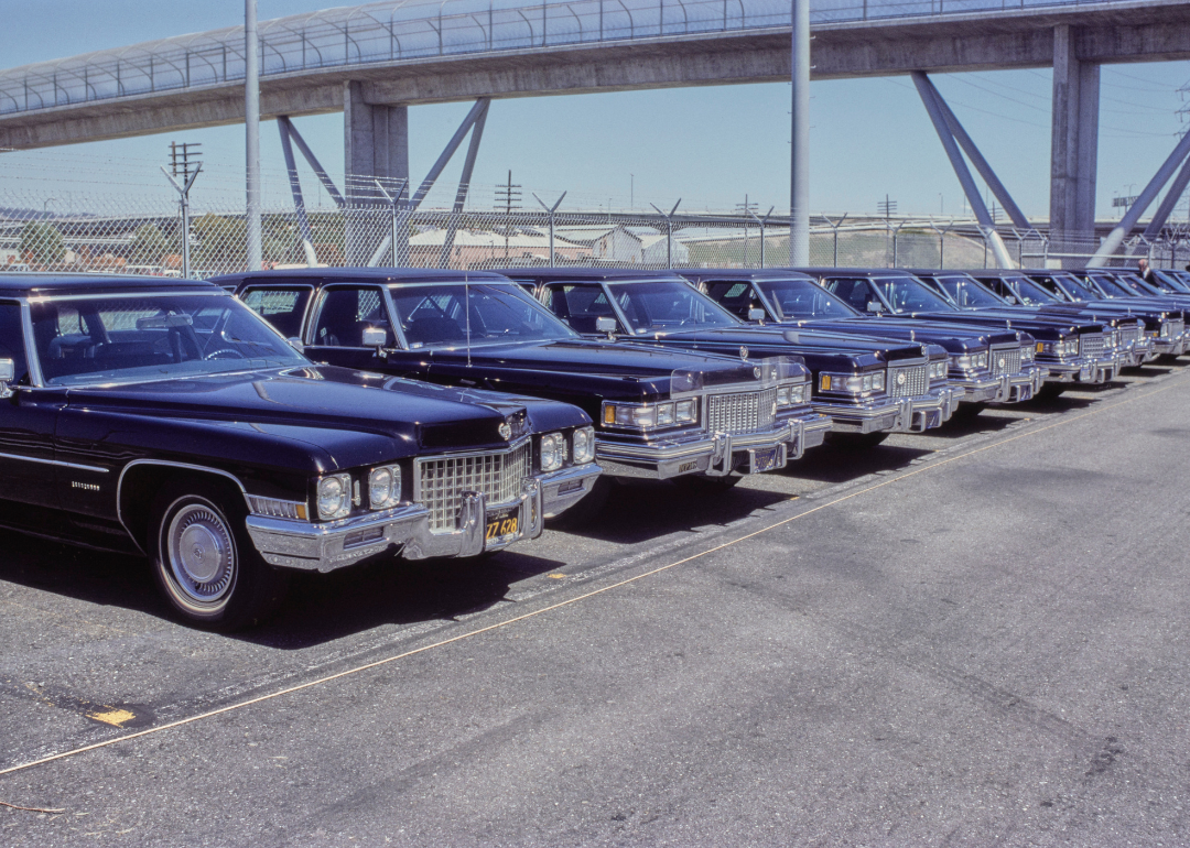 A Cadillac Eldorado (left) alongside a Cadillac DeVille (second left), among a line of various models of Cadillac car, all black, in a parked lot in May 1977. 