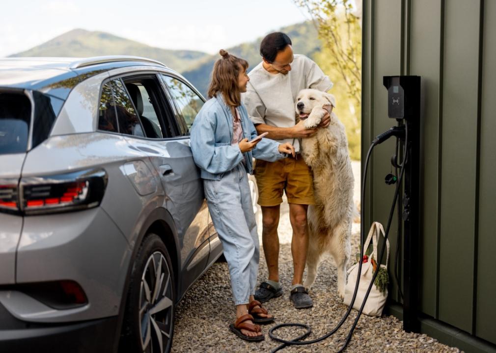 A young couple is outdoors charging the electric car. The man is hugging a large white dog, while the woman is watching them both.