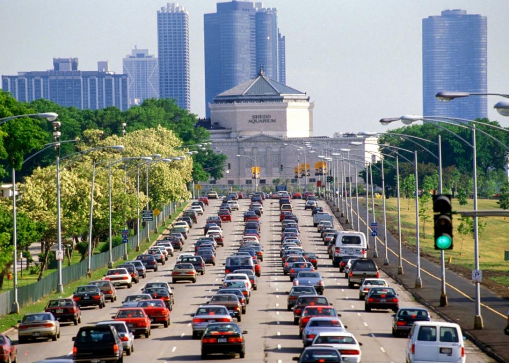 A multi-lane road lined with trees and streetlights. The road is filled with cars, creating a traffic jam. In the background, there are several tall buildings.