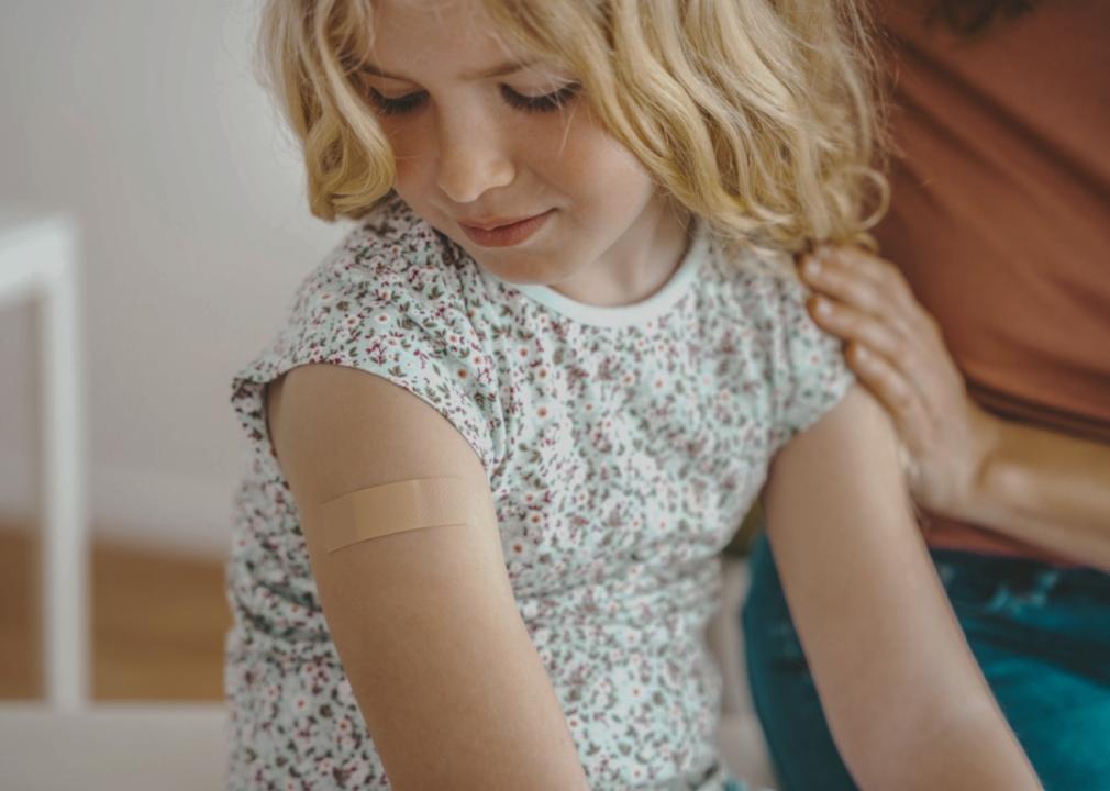 A young child with a small adhesive bandage on their upper arm. The child is wearing a floral-patterned shirt and looking down at the bandage.