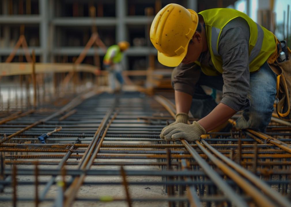 Construction worker wearing a yellow hard hat and safety vest, kneeling and working on steel bar at a construction site.