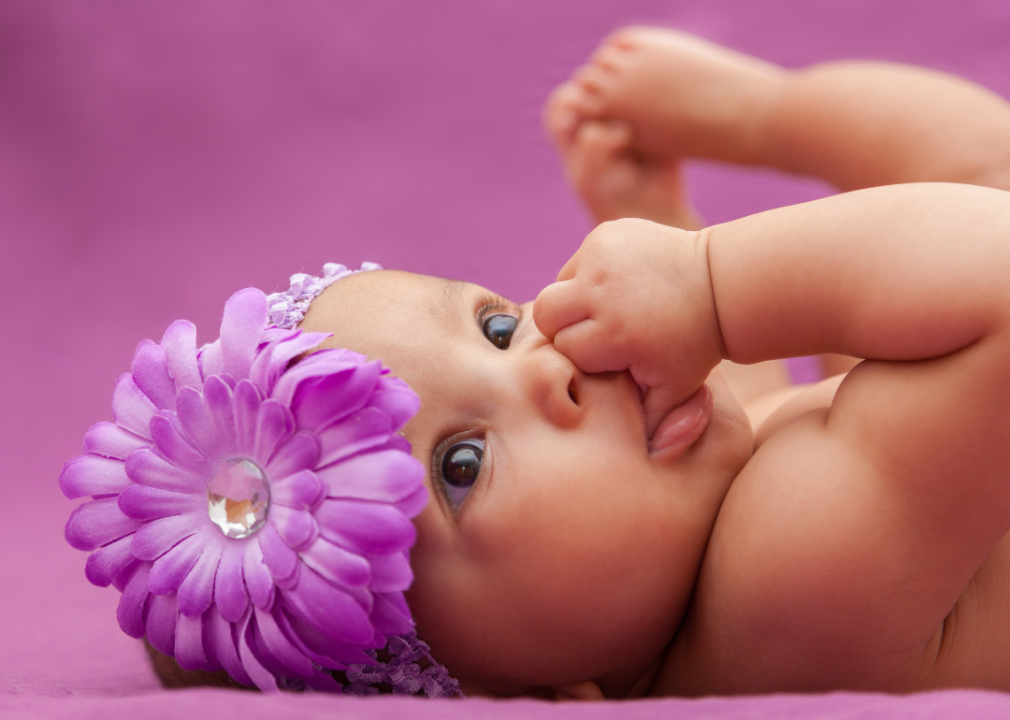 African American baby girl with a big purple flower on her head sucking her thumb.
