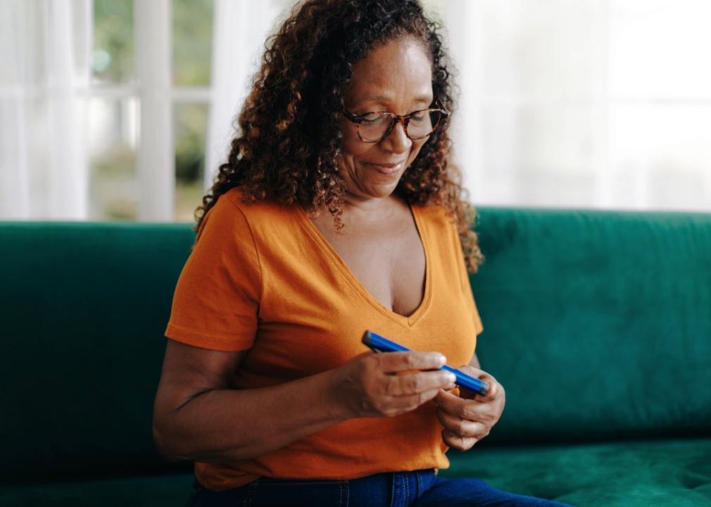 A woman sitting on a green couch, holding and looking at a blue object in her hands. The individual is wearing glasses and an orange shirt with a V-neckline. She is focused on the object they are holding.