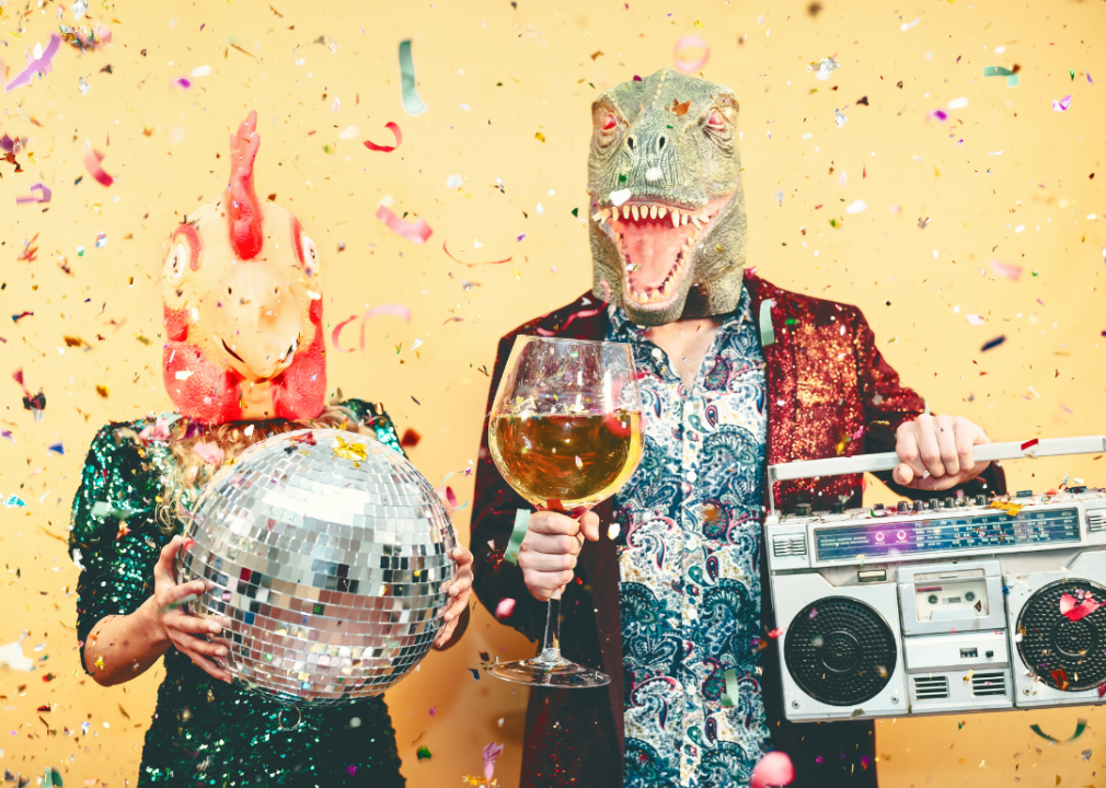 A woman wearing a rubber chicken head and a man wearing an alligator rubber head holding a disco ball, tape recorder and a large glass of wine.