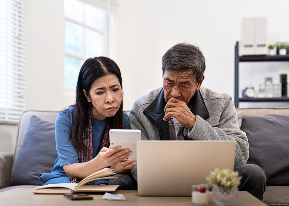 An elderly Asian couple is looking worried while sitting on a couch. There is a laptop on a coffee table, and the woman is holding a white square device, showing it to the man next to her.