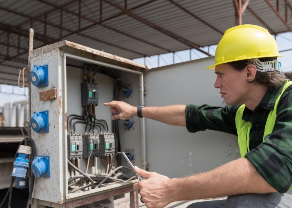 A man, likely an electrical technician wearing a yellow hard hat and a high-visibility safety vest. He is working with an open electrical control panel. The panel has multiple circuit breakers and wiring inside.