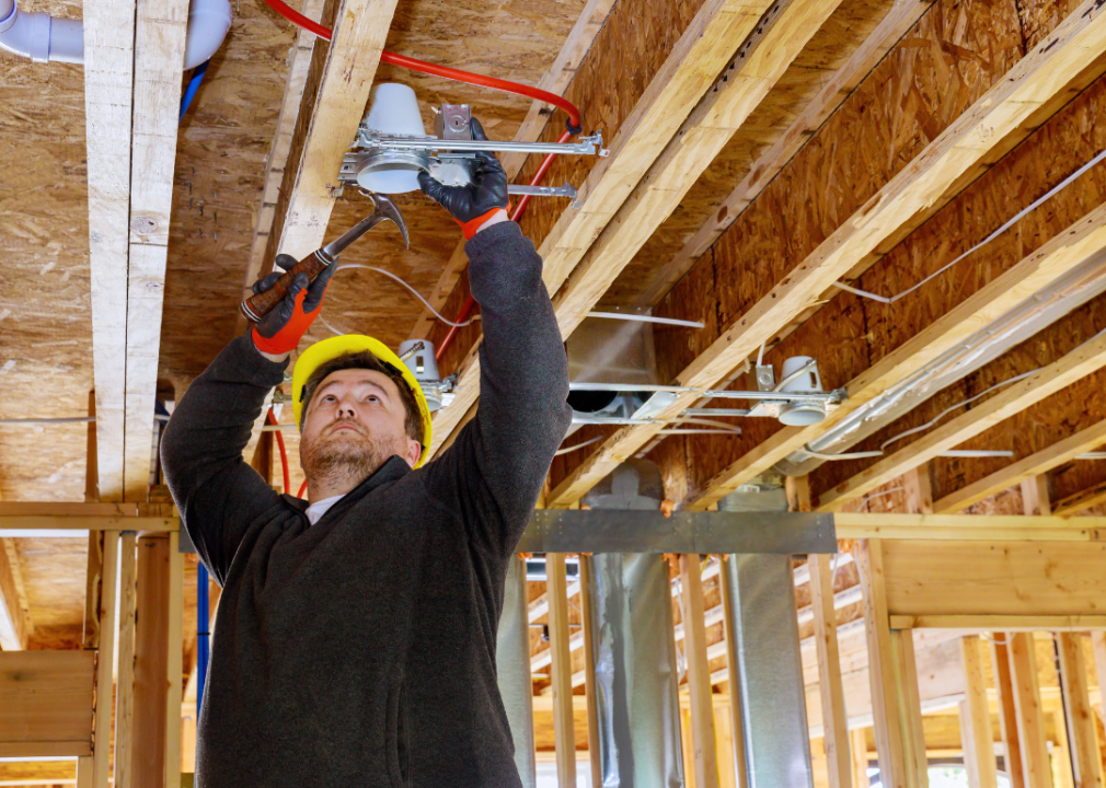 A man holds a light fixture in one hand and a hammer in the other in an unfinished construction place. 
