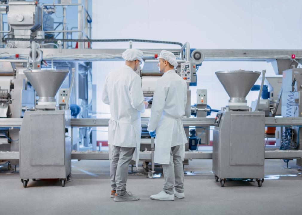 Two men dressed in white work robes and white hats standing in front of the equipment in a factory.