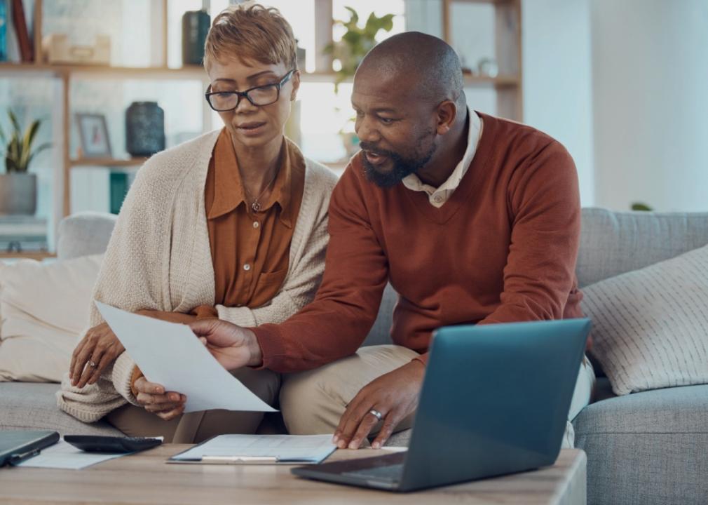 A couple sitting together on a couch in a cozy living room, reviewing documents. The woman is holding a sheet of paper and looking at it attentively. The man is pointing at the document. A laptop is open on the table in front of them, along with other papers and a calculator.