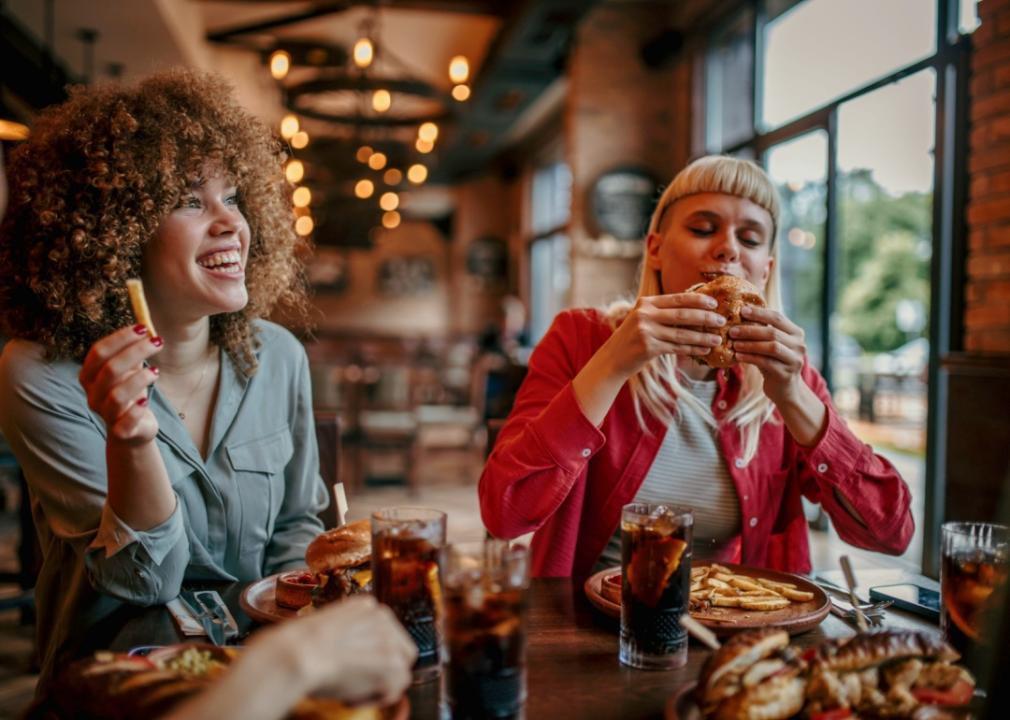 Two twenty-something women sitting at a table in restaurant eating burgers and fries.