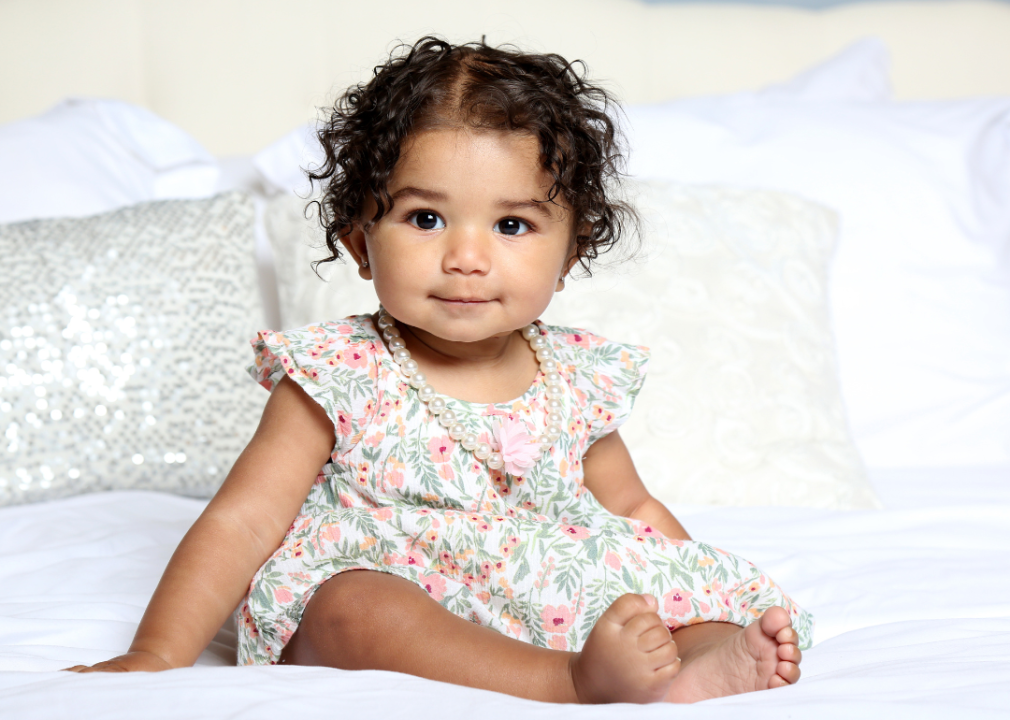 Hispanic baby girl with dark, curly hair in a flowery dress wearing pearls. 