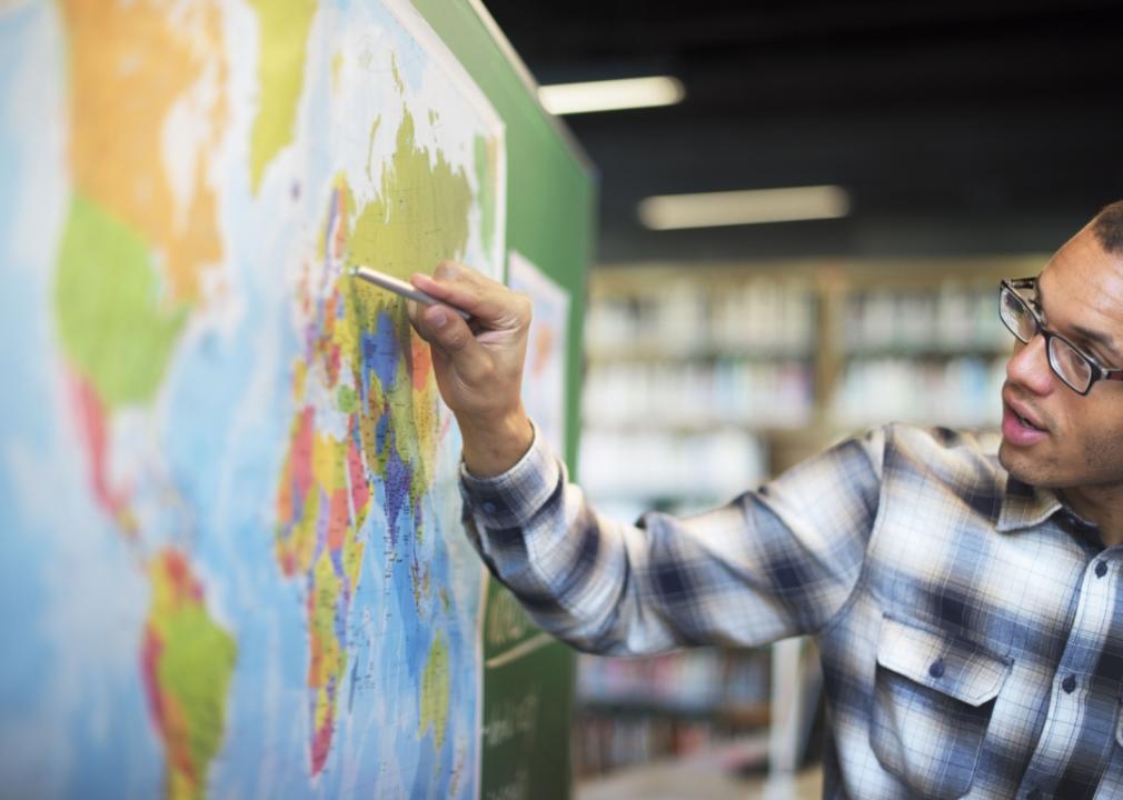 A young African American teacher with glasses is pointing at the world map in front of him. 