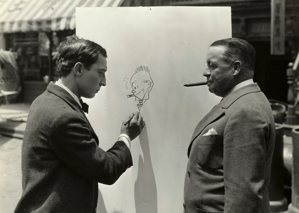 Buster Keaton pretends to work on a drawing actually started by cartoonist George McManus (right), who poses so as to mimic his character Jiggs from the comic strip Bringing Up Father.