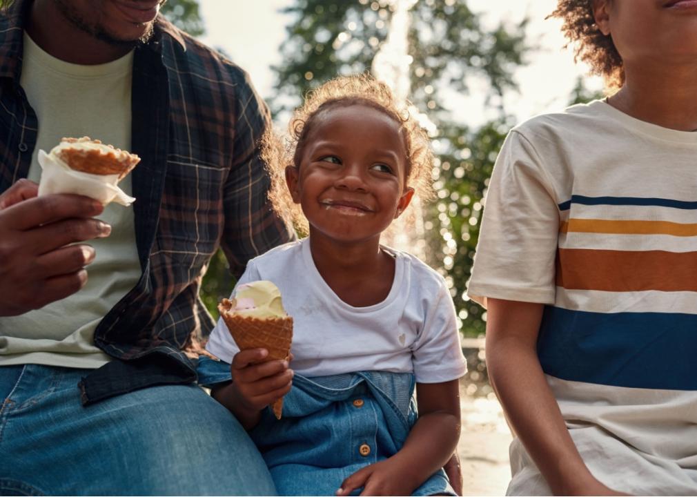 A joyful young black girl is holding an ice cream cone, seated between an adult man and a boy. 