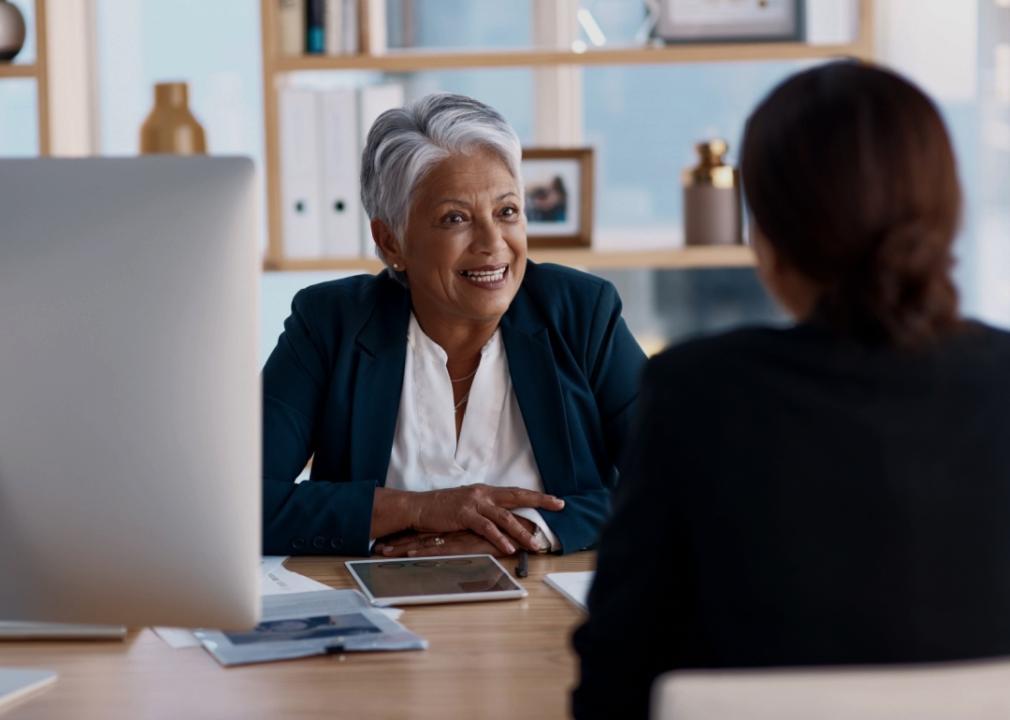 An older woman with short silver hair is looking at the woman sitting opposite her. She is wearing a dark blazer over a white blouse. She sits at a desk with various office items like a tablet, papers, and a desktop computer visible. On the right, there is another individual seen from behind.