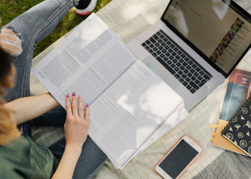 A student seated on a blanket in on the grass with an open textbook and laptop on their lap.