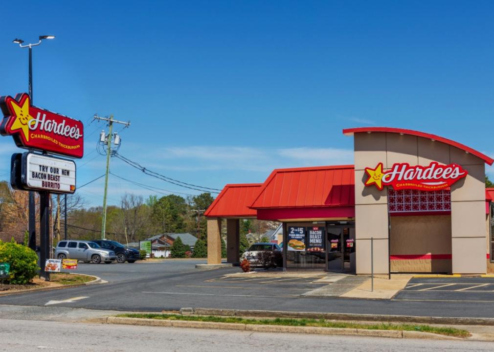 The outside of a Hardee's fast-food restaurant with a car stopped at the driv-thru window and a few other cars in the parking lot. 