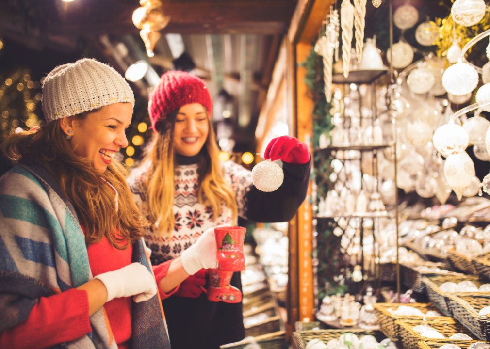 Two women smiling at the outdoor holiday market while looking at the Christmas tree decorations. 