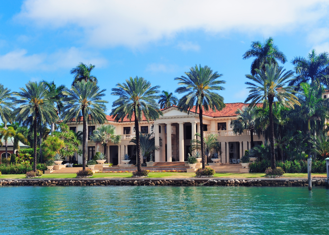 A luxury home with large columns in front and palm trees on the water.