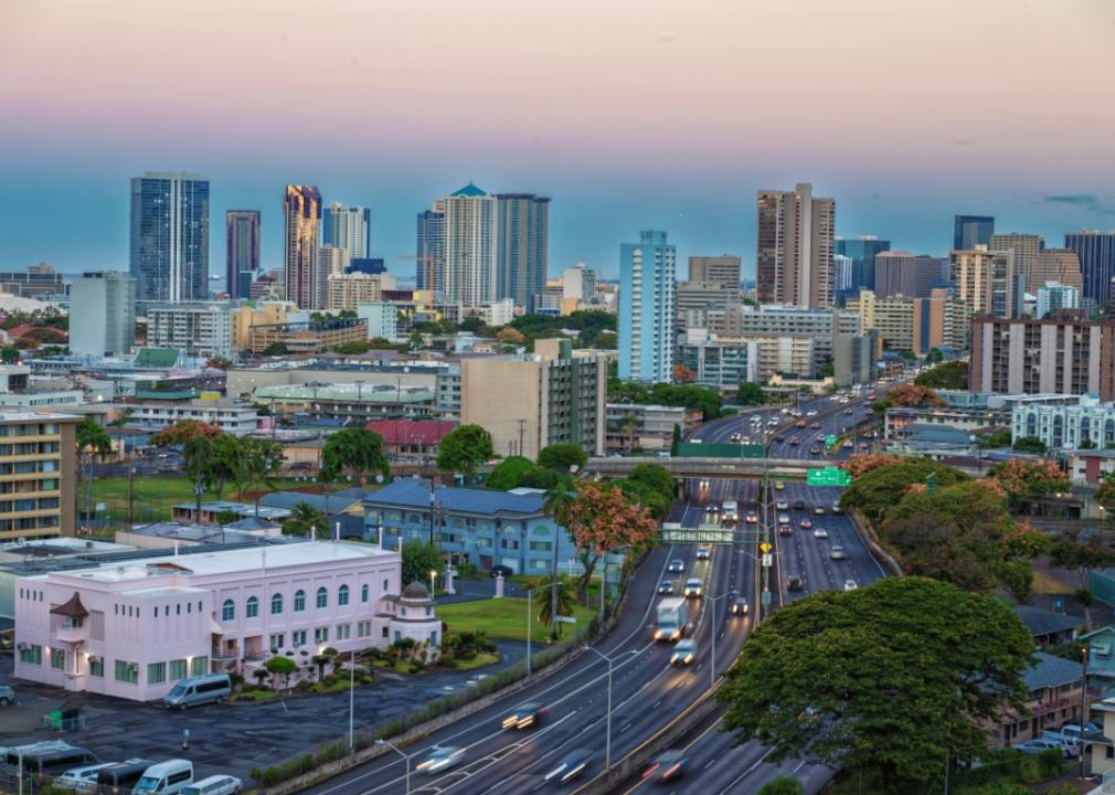 City skyline dominated by tall skyscrapers in the background. A wide, multi-lane highway cuts through the city, filled with traffic.