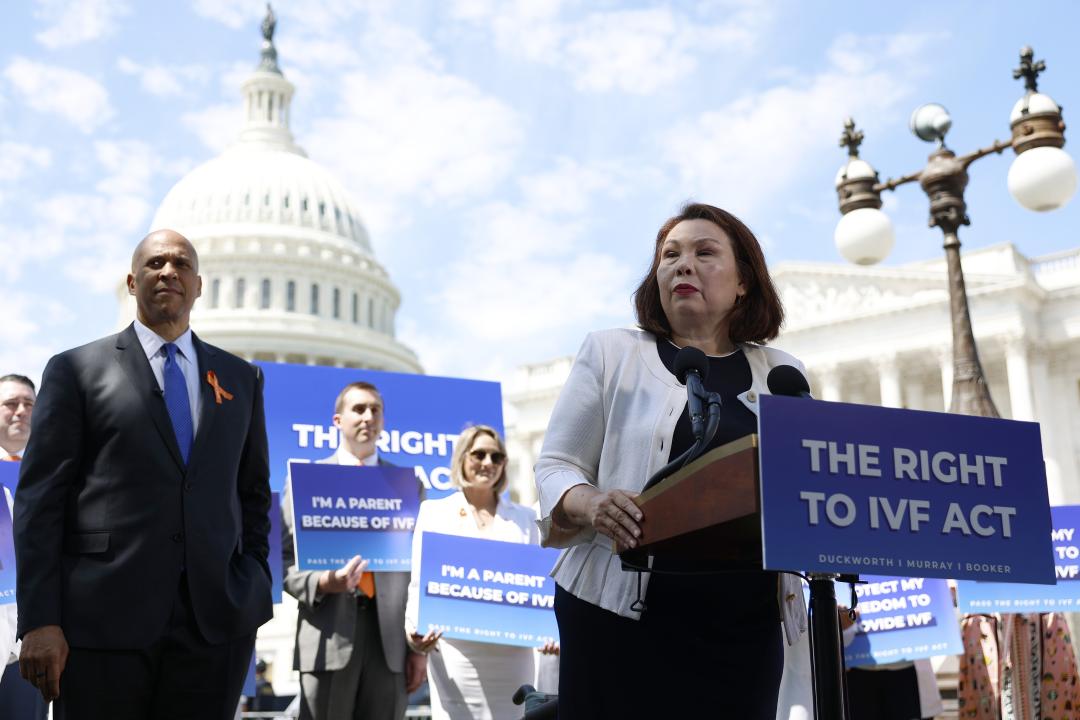 Senator Tammy Duckworth speaks alongside Senator Cory Booker during a news conference on access to IVF treatments outside of the U.S. Capitol Building. The podium in front of her has a poster that reads, The Right to IVF Act while people behind her hold signs that say, I