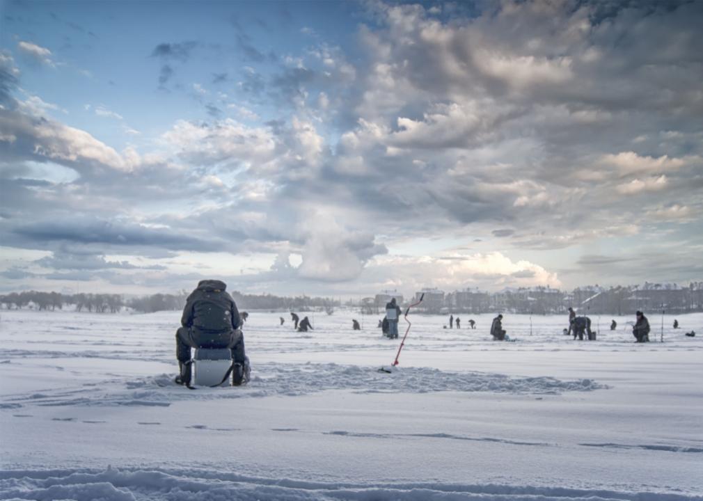 A person sitting on top of a cooler while ice fishing in the foreground with more people ice fishing in the background. 