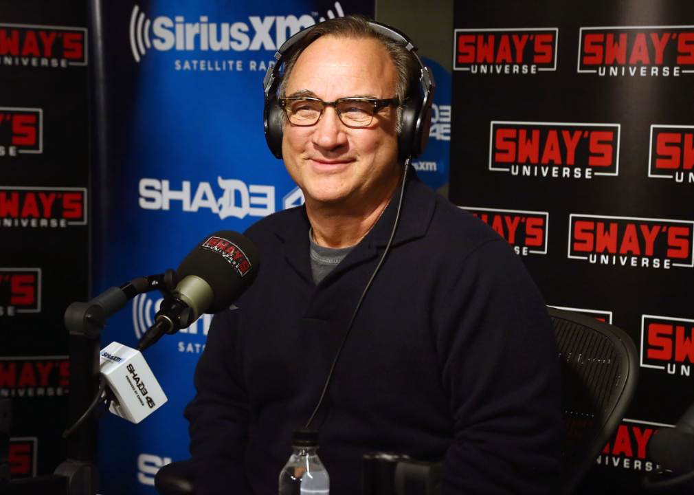 Jim Belushi sitting in front of a mic with headphones on, with SiriusXM Satellite Radio logos and Sway's Universe logos on posters behind him.