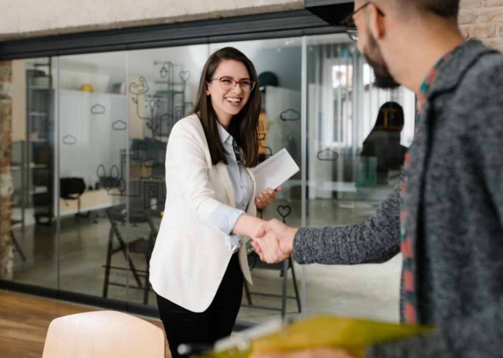 A young woman smiling, shaking man's hand in an office space.