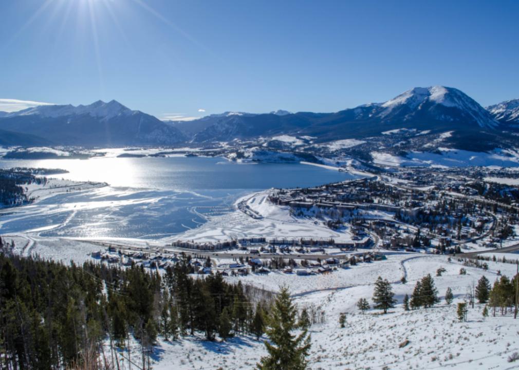 An aerial view of a vast partially frozen lake and town surrounded by tall snowy mountains. 