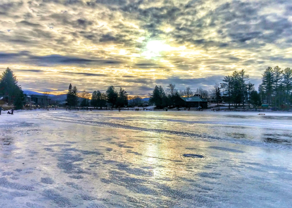 People ice skating on a frozen lake surrounded by tall trees and mountains in the background. 