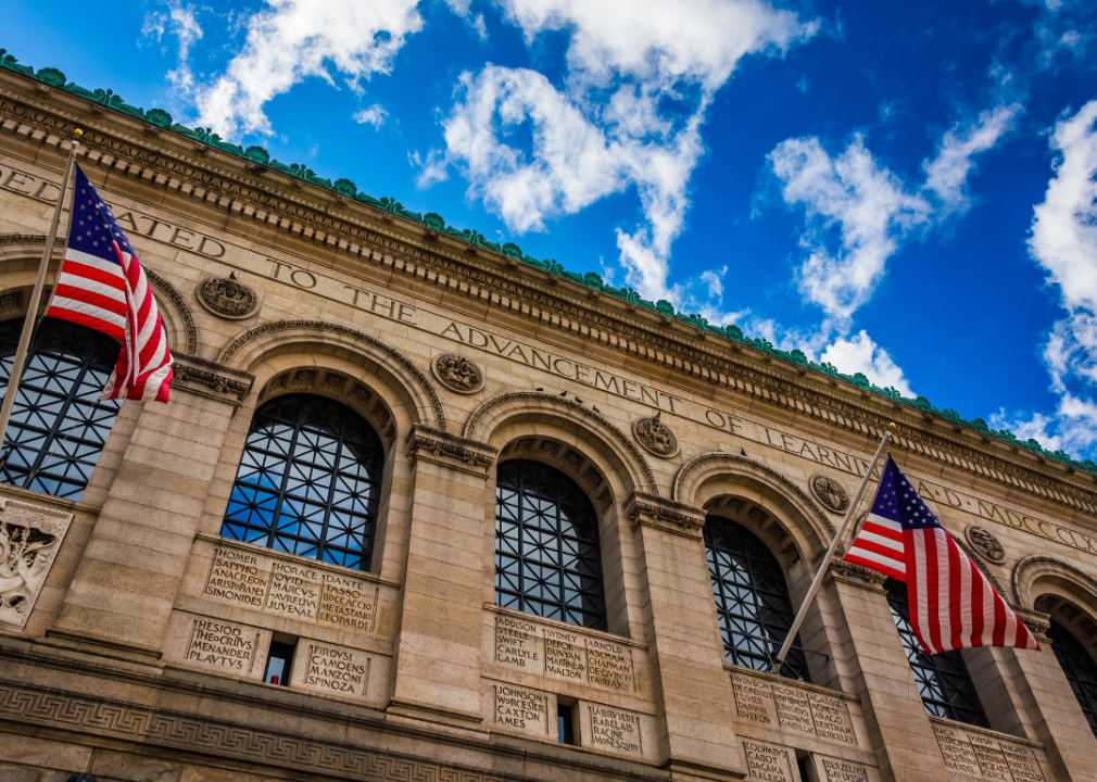 A closeup of part of a building facade made of light colored stone. Two American flags flying in front of the building on flagpoles. 