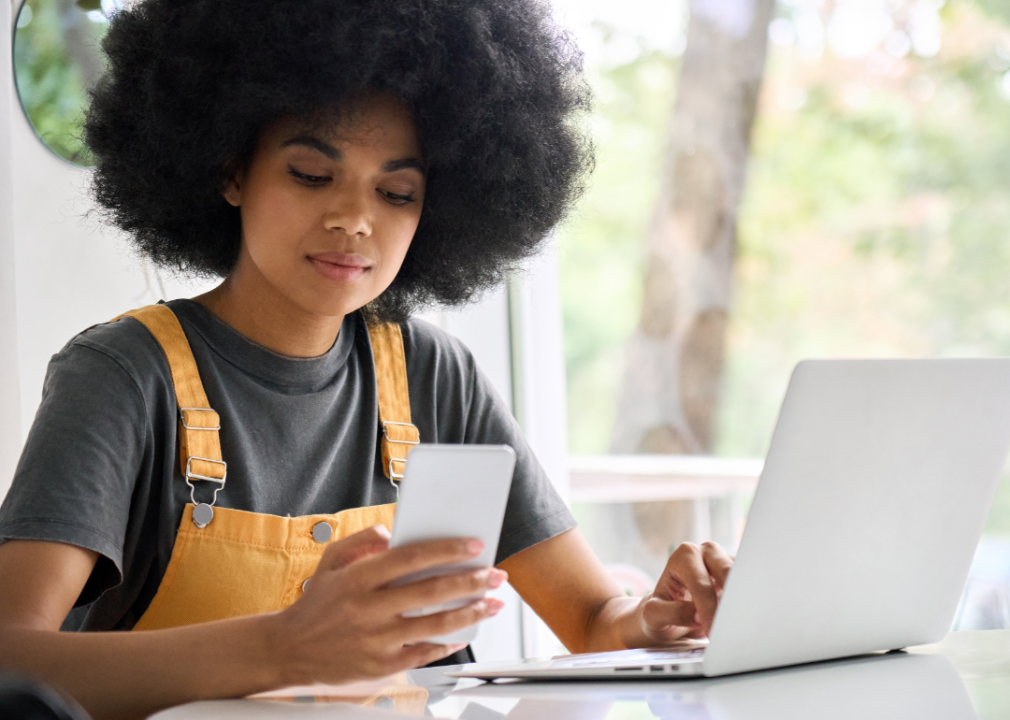 A young woman simultaneously checks her phone and her laptop