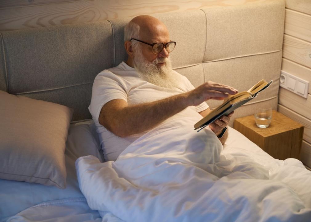 A bearded older man wearing glasses lies in bed while holding a book.