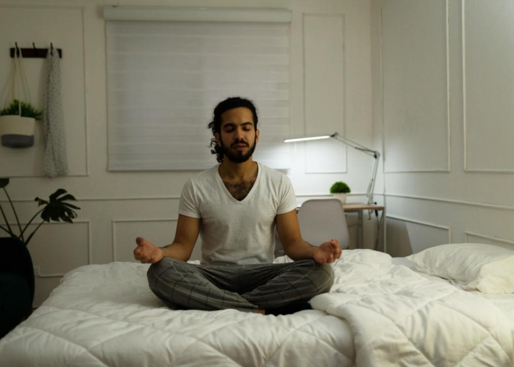 A man in pajamas sits cross-legged in bed with his eyes closed in meditation.