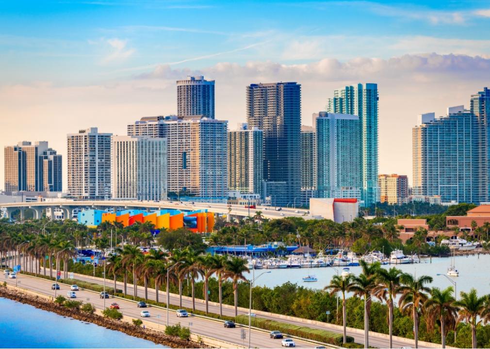 A coastal city with a cluster of modern high-rise buildings along the waterfront. A busy highway runs alongside the coast, lined with palm trees.