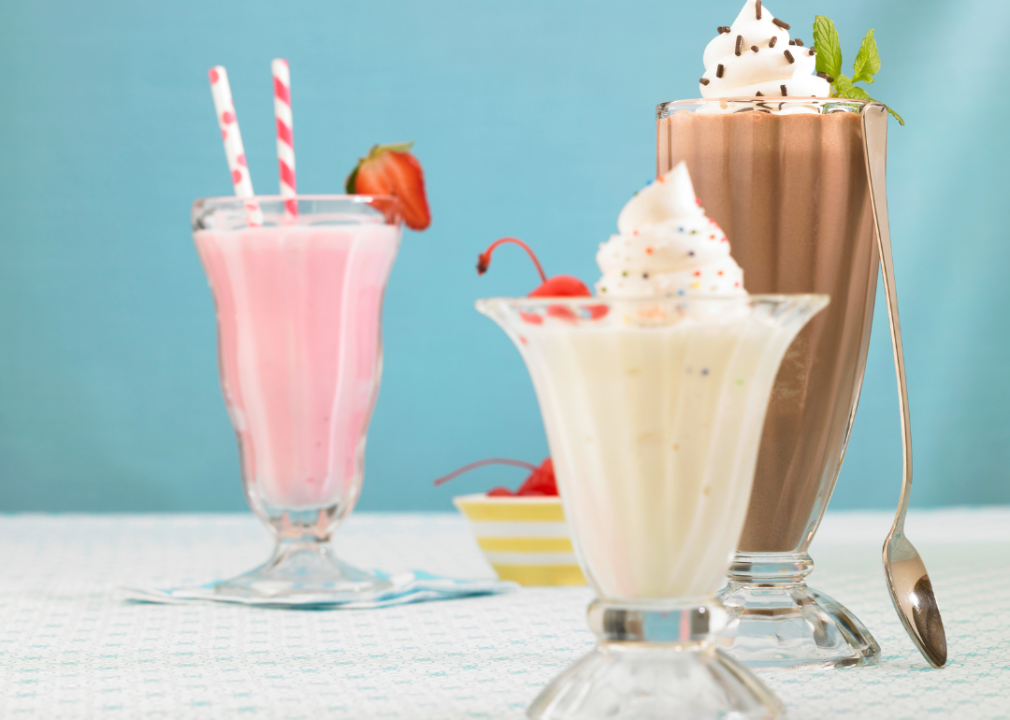 Three milkshakes topped with whipped cream in tall glasses: the one on the left is light pink, the one in the middle is light brown, and the one on the right is light chocolate brown. 