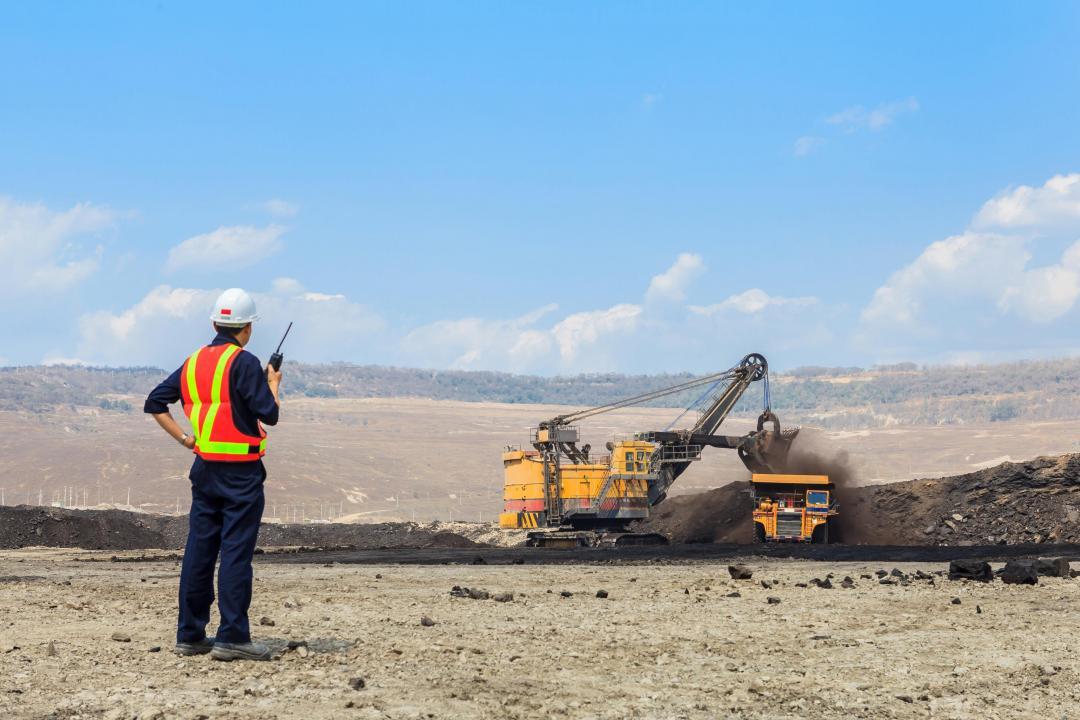 In a wide open field, a man in a hard hat and high-viz vest holds a walkie talkie and supervises large machinery being operated.
