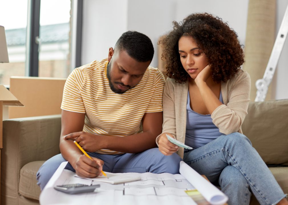 A concerned young couple reviewing their finances while looking at a blueprint for their home.