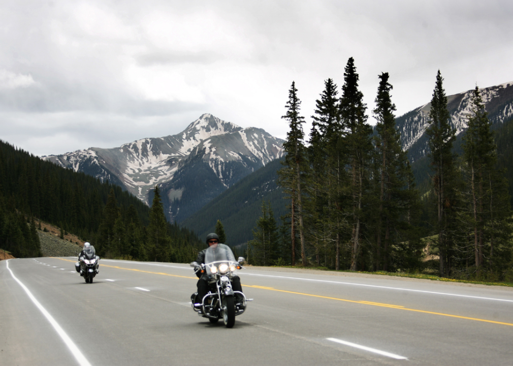 Two people riding motorcycles on a highway with the Rocky Mountains in the background.