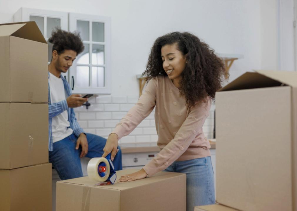 A young couple in a kitchen surrounded by boxes. A woman is holding a packing tape and taping a large box on the floor while a man is sitting down on the counter next to a large pile of boxes looking at a phone in his hand. 