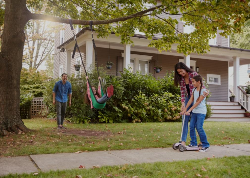 A family in front of a suburban home with a well-maintained lawn and a tree with a swing. A young girl is swinging while a man stands nearby with a smile, seemingly pushing. To the right, a woman is helping another young girl ride a scooter on the sidewalk. 