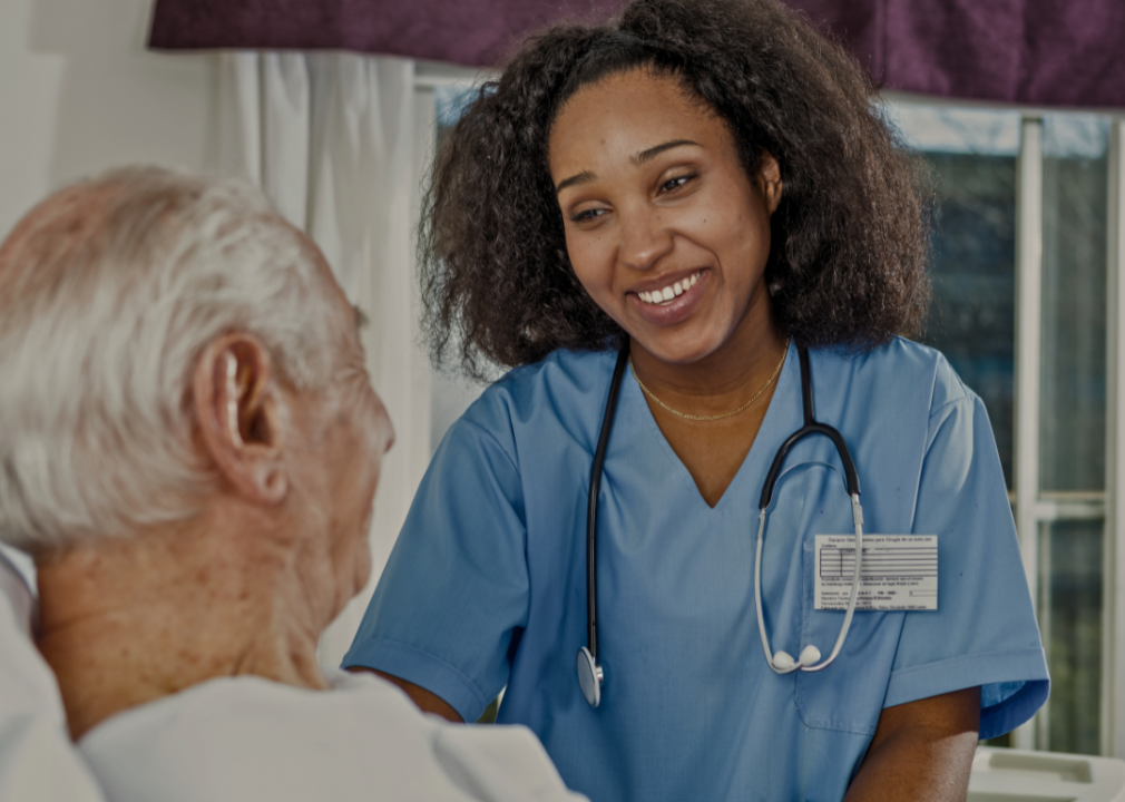 A nurse wearing blue scrubs and a stethoscope around her neck. She is smiling warmly at the patient, who is facing her and sitting up in a hospital bed.